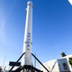 A part of the Falcon 9 Rocket with the SpaceX logo stands up on a large tripod surrounded by nearby buildings